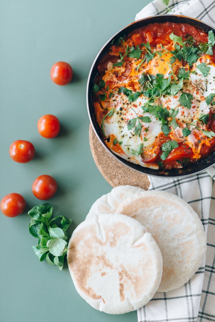 Top view of shakshuka with pita bread on a green table, garnished with herbs.